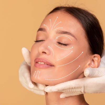 Beauty treatment concept. Young woman receiving face massage from cosmetologist, doctor wearing protective gloves, beige studio background, cropped